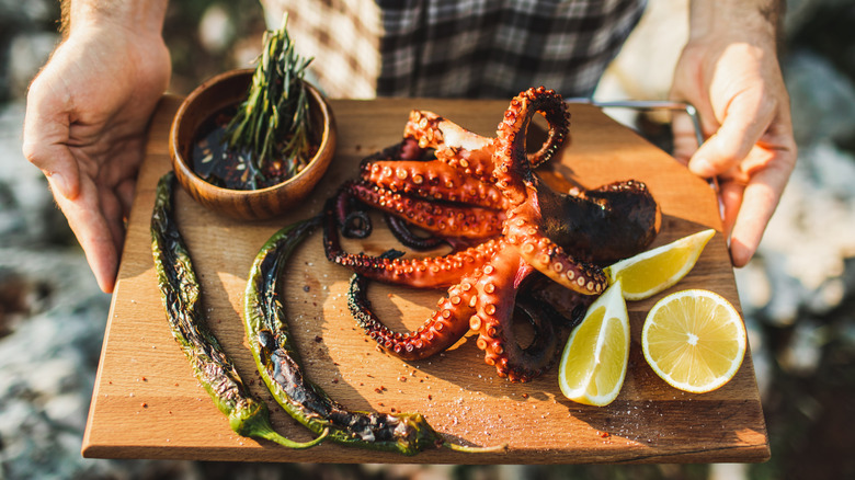 Vibrant octopus served on a wooden board with charred peppers, herbs and slices lemons.