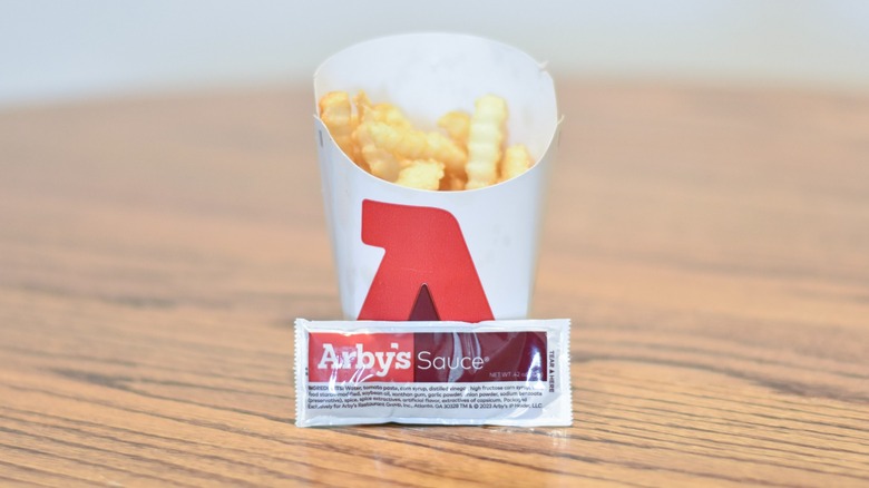 A packet of Arby's sauce in front of a carton of fries on a wooden table