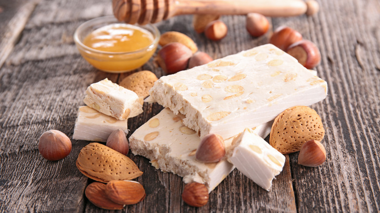 Bars of white nougat are placed on a rustic wooden table surrounded by honey and nuts.