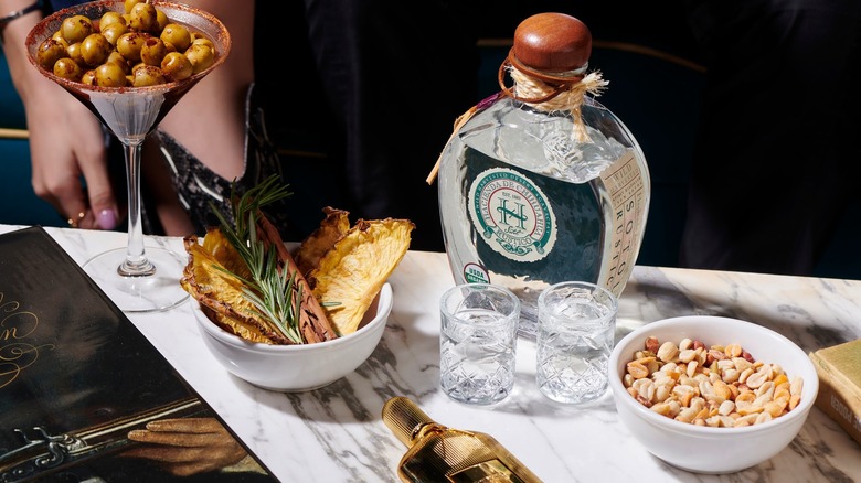 A bottle of Hacienda de Chihuahua with shot glasses, peanuts, dried fruit, and a martini glass of olives
