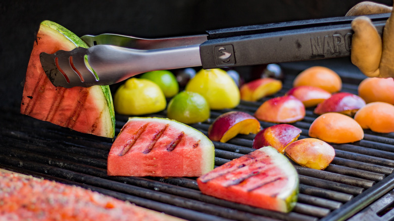 Tongs flipping watermelon on a grill with limes, lemons, and nectarines grilling in the background