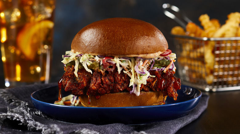 Nashville chicken sandwich with slaw rests on a blue plate with a basket of fries nearby.