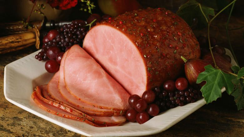 A partially sliced, whole glazed ham on a platter surrounded by grapes on the vine and figs, a carving fork and knife in the background.