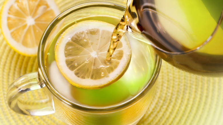 Pouring hot tea into a glass mug with a lemon wheel floating in the tea, a sliced lemon to the side