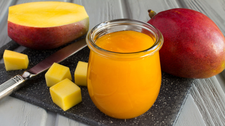 A small bowl of mango puree sits beside cubed mango pieces, as well as sliced and whole mangos