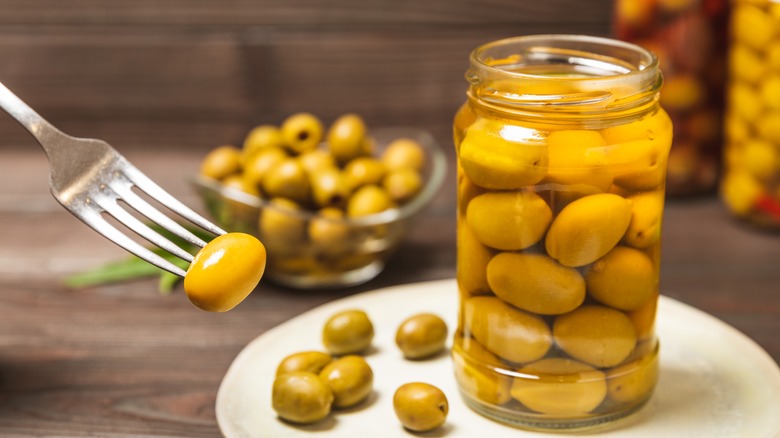 A jar of green olives in brine next to a single olive on a fork