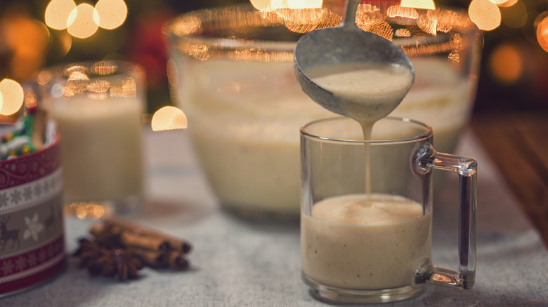 Creamy eggnog being ladled into a glass, with a bowl of eggnog and cinnamon sticks in the background.