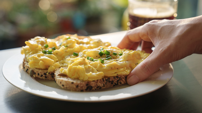 Hand picking up fluffy scrambled eggs on toast served on a white plate