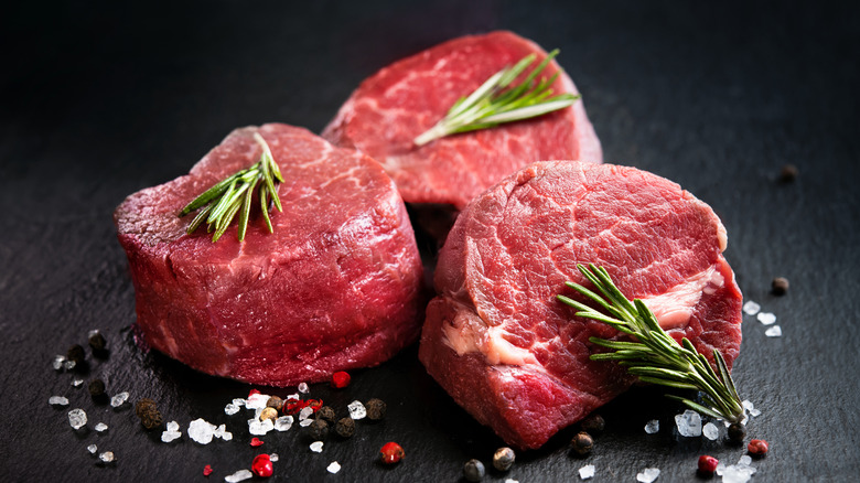 Three uncooked cuts of filet mignon with rosemary sprigs, salt, and peppercorns.