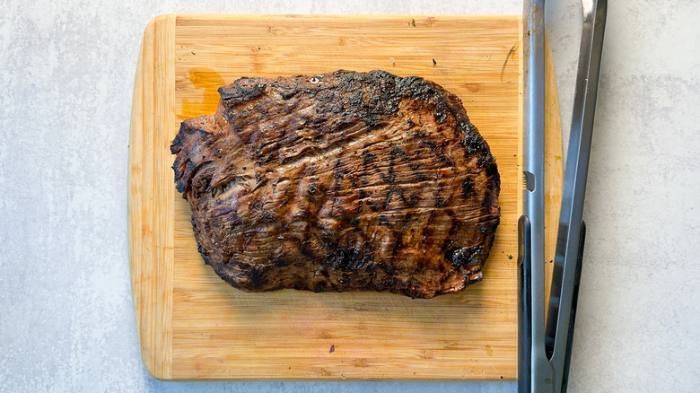 Grilled flank steak resting on carving board with tongs