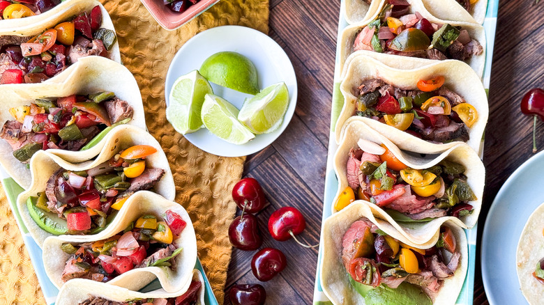 Grilled flank steak tacos with stone fruit salsa on serving platters on table with limes and cherries