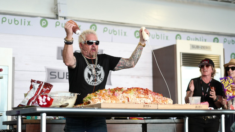 Celebrity chef Guy Fieri squeezes sauce onto a huge tray of food at a live cooking demonstration.