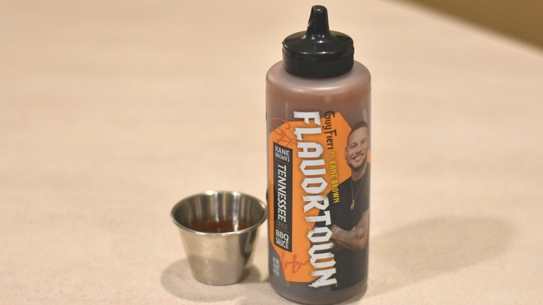 A bottle of Flavortown Kane Brown Tennessee barbecue sauce next to a small silver bowl with a sample of dark red sauce