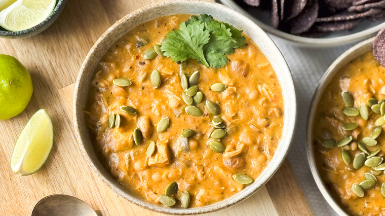 Hearty white chicken and navy bean chili in bowls with blue corn tortilla chips, pepitas, and lime wedges