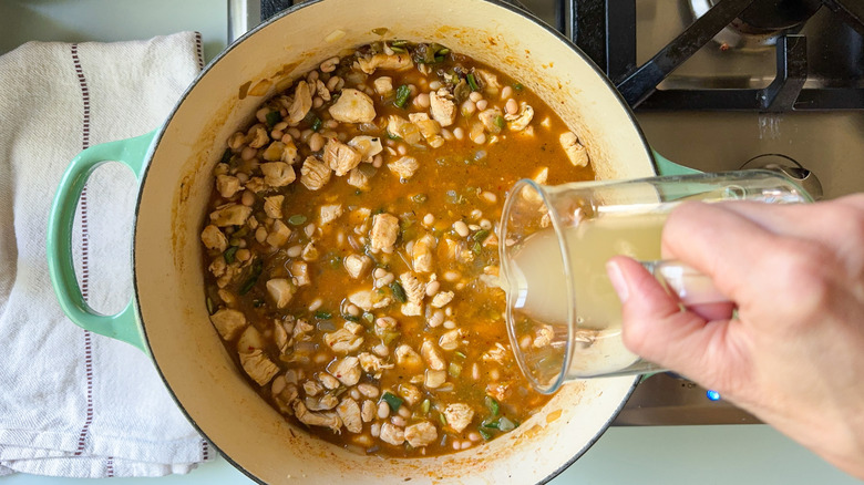 Pouring chicken broth into white chicken and navy bean chili in large pot on stovetop