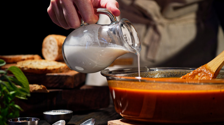 Closeup of a hand pouring a pitcher of heavy cream into a bowl of tomato soup