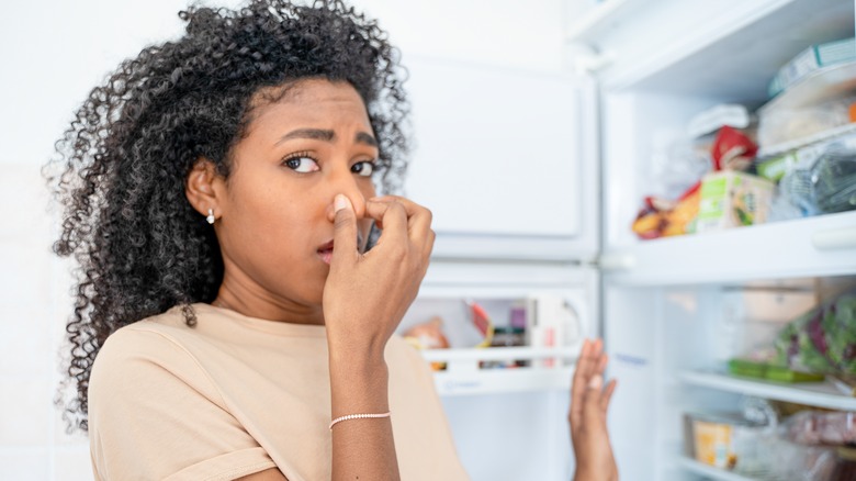 Woman holding nose next to open fridge. 