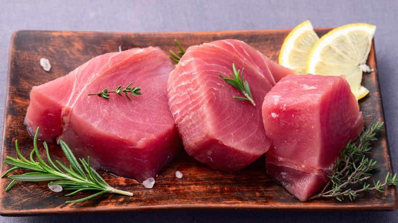 Three fresh, thick tuna steaks on a wooden platter, surrounded by thyme and rosemary sprigs, fat salt crystals, and two lemon wedges, topped with smaller sprigs of thyme and rosemary.