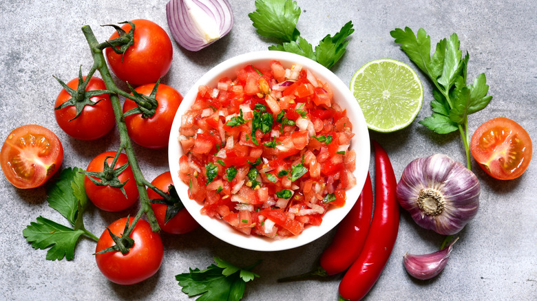 A bowl of mostly tomato salsa with tomatoes on the vine, bulbs of garlic, lime and cilantro surrounding.