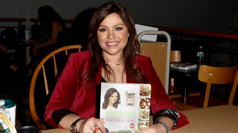 Rachael Ray sitting at a table holding one of her cookbooks