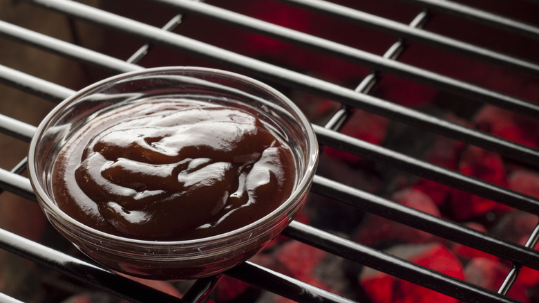 A bowl of barbecue sauce sits atop a grill in close up.