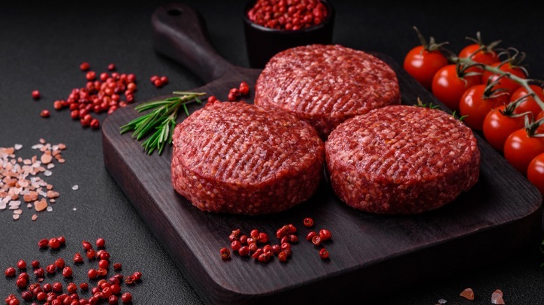 Three raw burger patties, with spices and tomatoes around