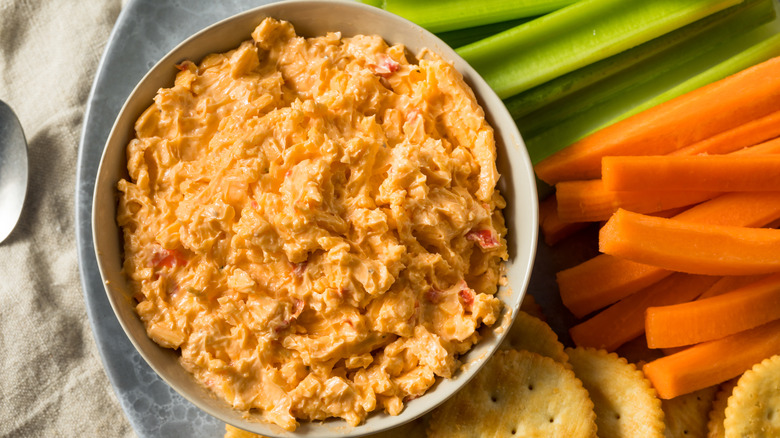 Pimento cheese dip is served in a white bowl alongside crackers and fresh veggie sticks.