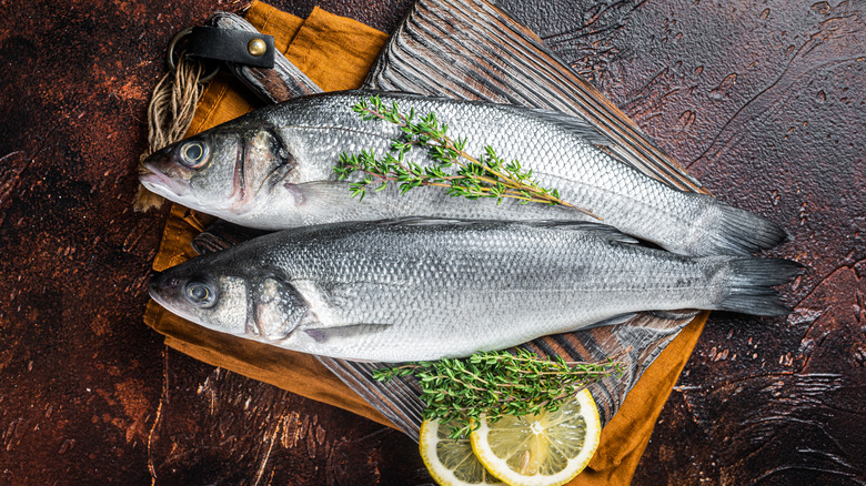 Two silver fish laying on a wooden cutting board with springs of green herbs and slices of lemon, all sitting on an orange towel