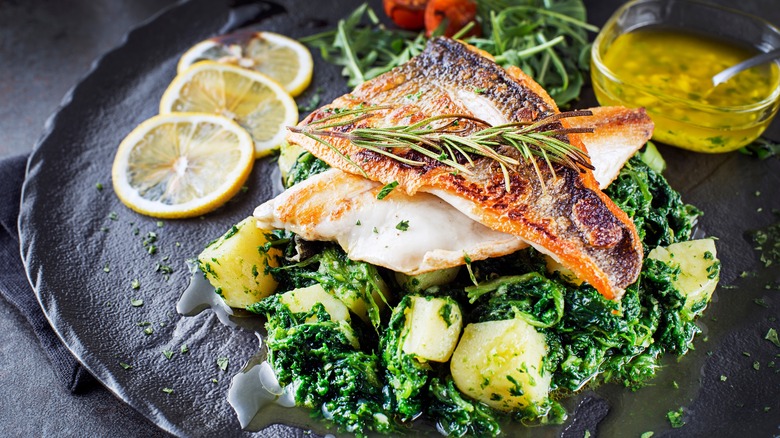 Fried filets of white fish sitting atop cooked greens and potatoes with a glass bowl of oil with a spoon in the background and three slices of lemon, all sitting on a black plate