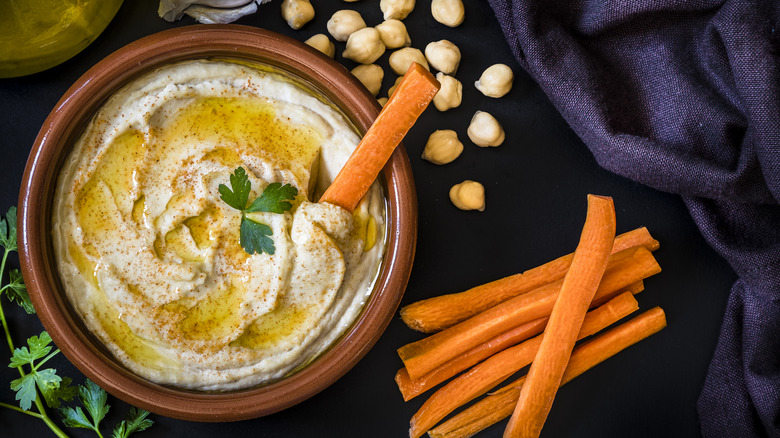 Bowl of hummus with carrot sticks and chickpeas on dark purple background