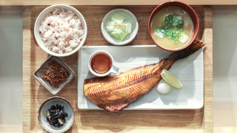 wooden tray holding bowl of miso soup, white rice, cooked fish, dipping sauce, lemon wedge, and various vegetables