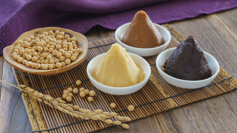 three different types of miso paste in white bowls on bamboo mat next to bowl with dried soybeans