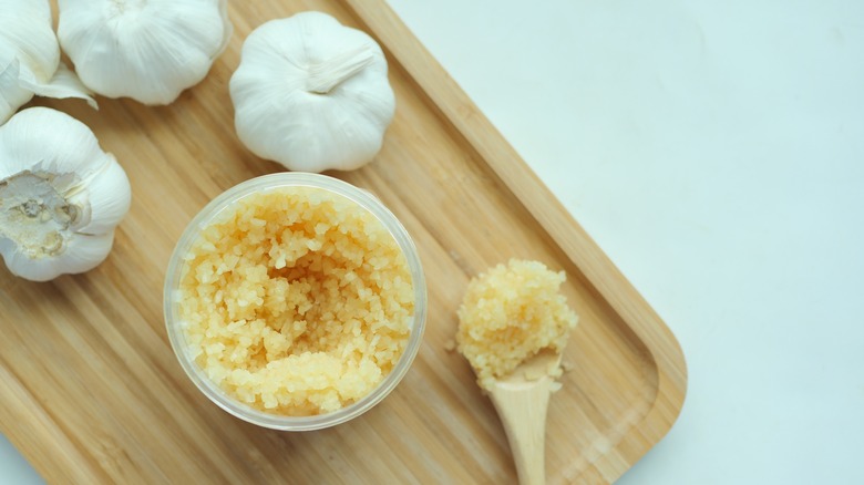 A bowl and spoon of minced garlic next to whole garlic bulbs on a wooden tray