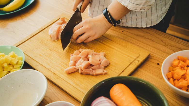 Overhead view of person's arms cutting chicken on wood cutting board into cubes