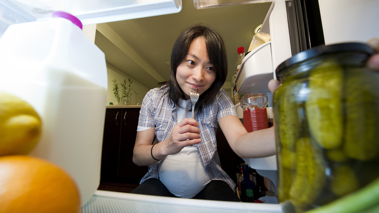 A woman grabs a jar of pickles in the refrigerator. 