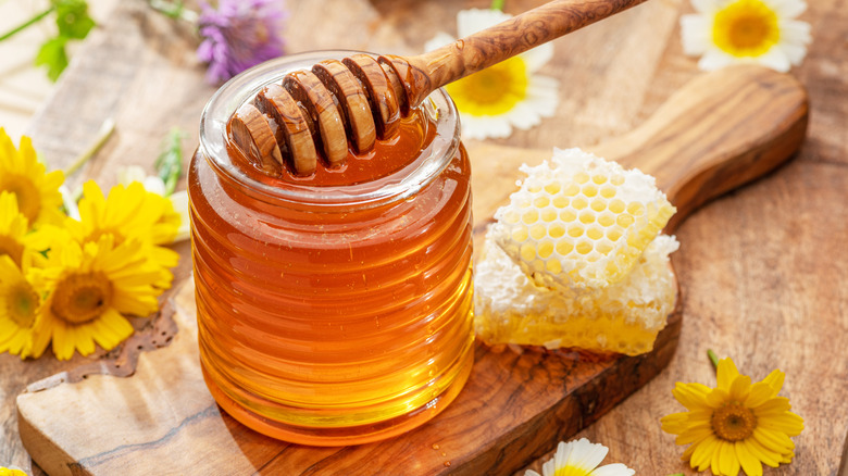 jar of honey with a stirrer surrounded by flowers and honeycomb