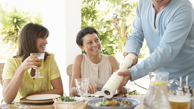 Man using a large pepper mill over a salad as two women eat and smile at the table.
