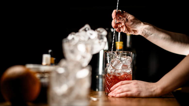 A bartender stirring a Sazerac. cocktail in a glass with cocktail accoutrements surrounding.