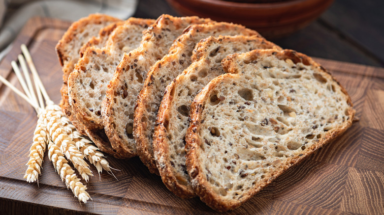 Crispy, airy sliced sourdough bread on a wooden cutting board with a bundle of wheat stalks next to them.