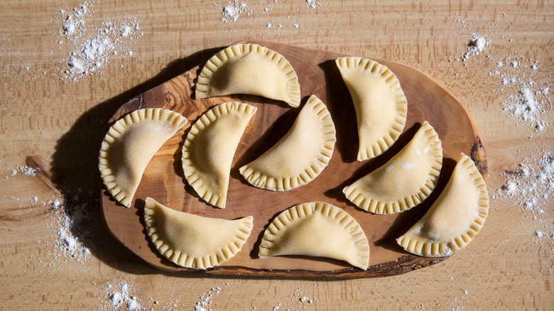 Uncooked ravioli sitting on a rustic wooden board on a flour-sprinkled table.