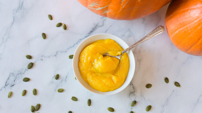 Pumpkin puree in bowl with spoon along with pepitas and pumpkins in background