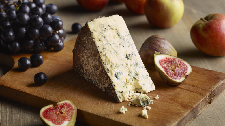 A wedge of blue cheese and some crumbles on a wooden cutting board with fruit