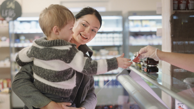 Mother holds child, who reaches out for a sample of deli meat