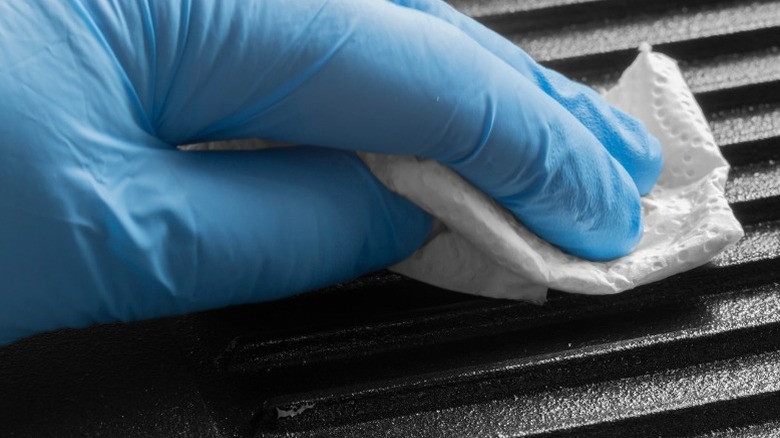 A gloved hand wipes a cast-iron grill pan with a paper towel.