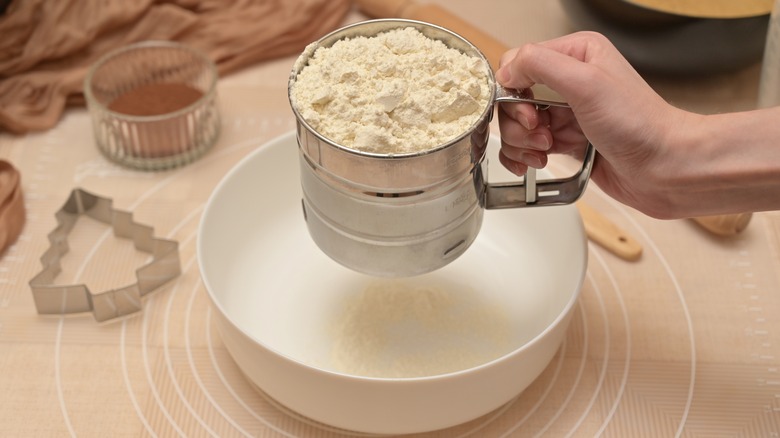 Person holding a sifter with flour over a bowl