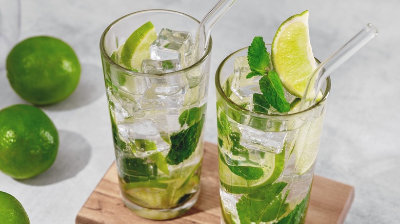 Two glasses of mojito sit on a wooden board surrounded by limes.