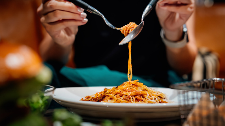 A person's hands twirl spaghetti with a knife and spoon over a large plate.