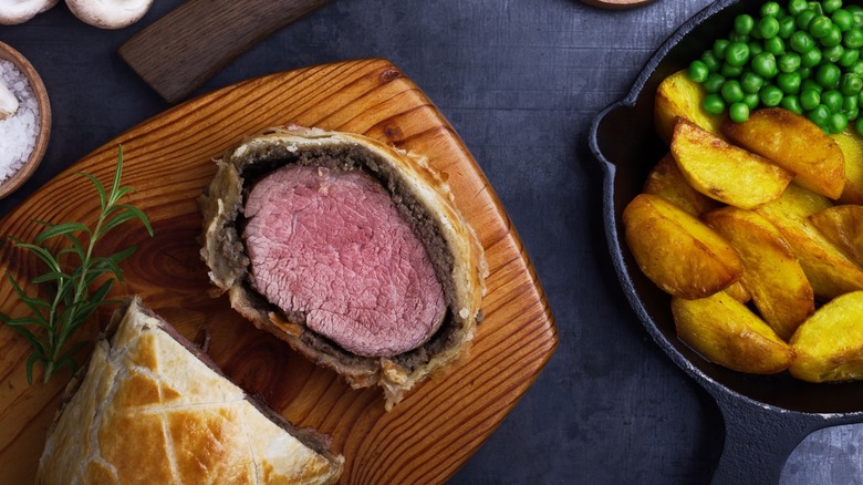 Cut Beef Wellington on wooden tray next to bowl of potatoes, peas