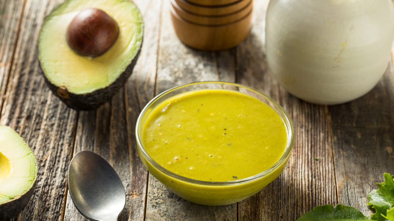 A bowl of green goddess salad dressing surrounded by two avocado halves, an olive oil carafe, a pepper grinder, spoon, and lettuce leaves.