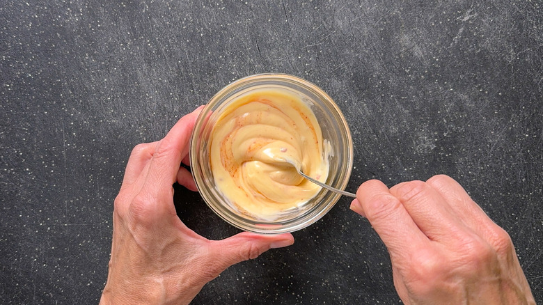 Mixing Kewpie mayo with chili sauce in small glass bowl with spoon on cutting board